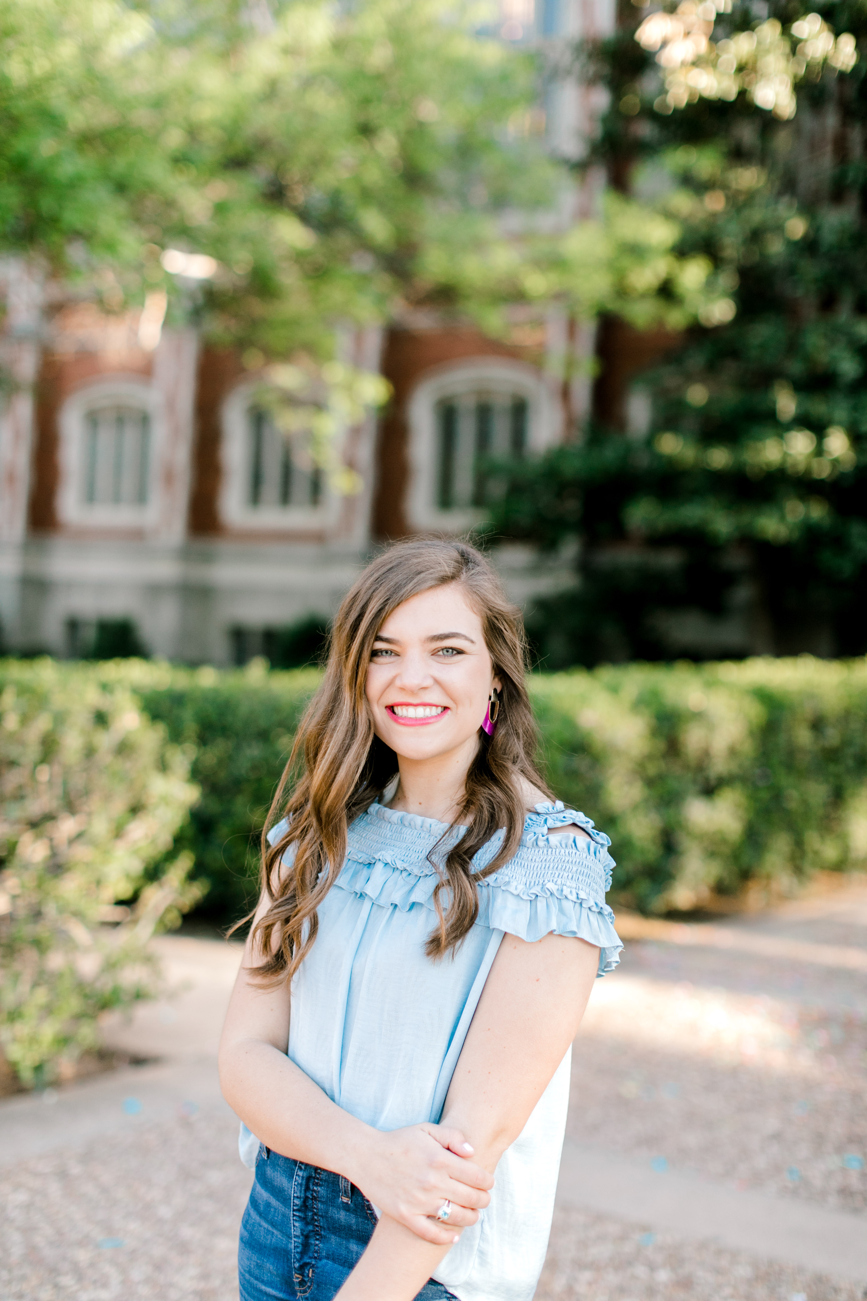 brunette color senior in blue blouse stands in front of collegiate building for OU graduation photographer