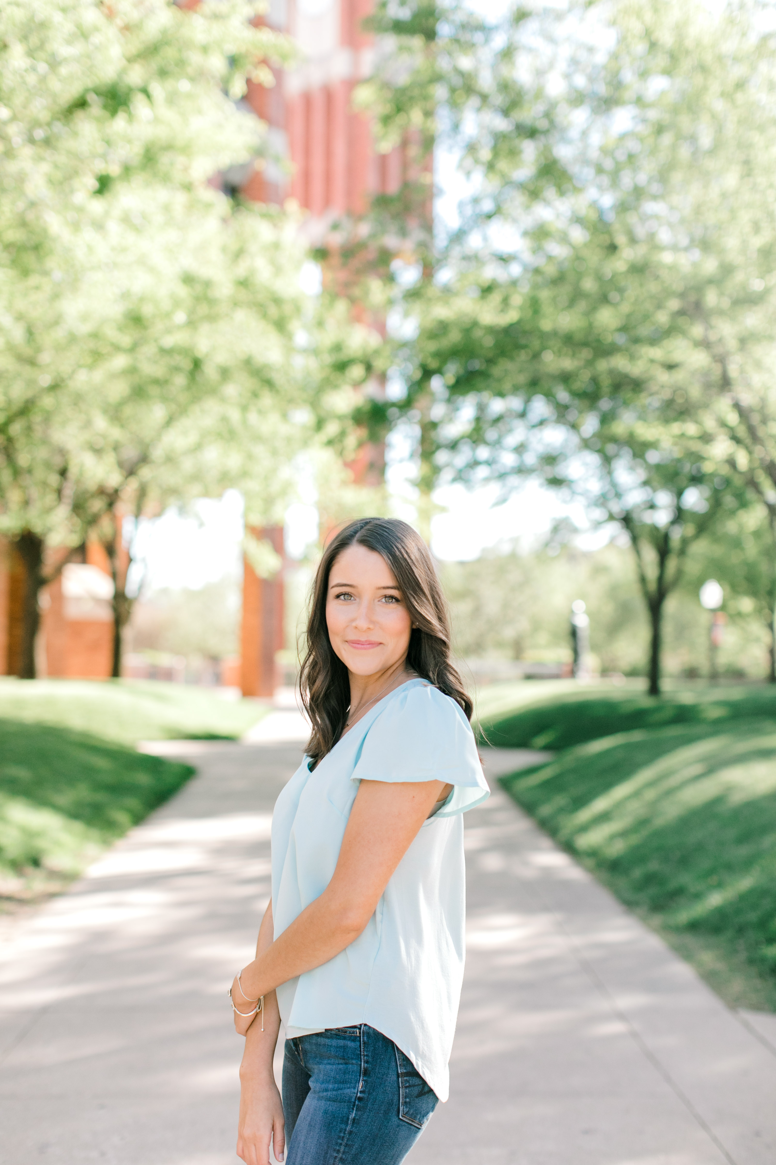 brunette girl in blue holds arm and poses in front of green trees on ou campus for ou graduation photographer