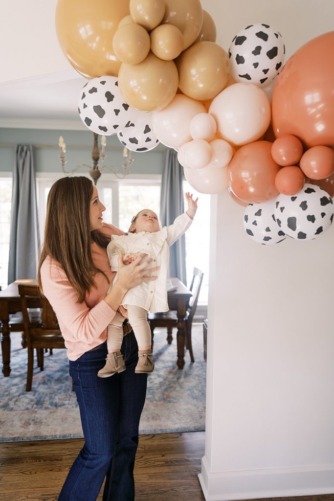 Mom holds 1 year old baby for first birthday party under cow printed balloon arch